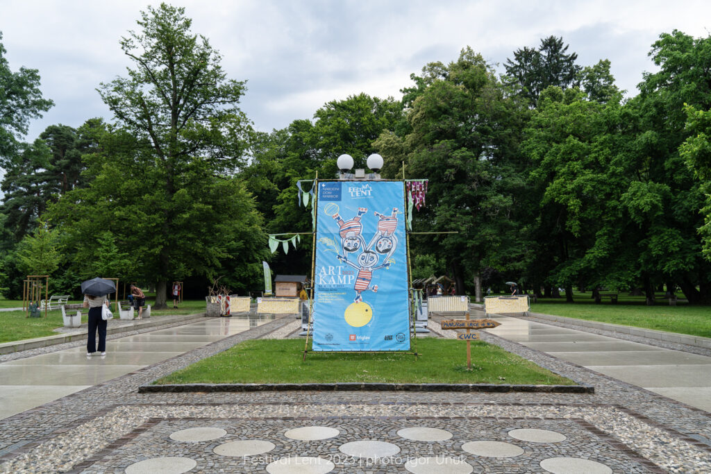 Prizorišče dogajanja, velik pano z logotipom Artkampa na zelenici na promenadi Mestnega parka v Mariboru.