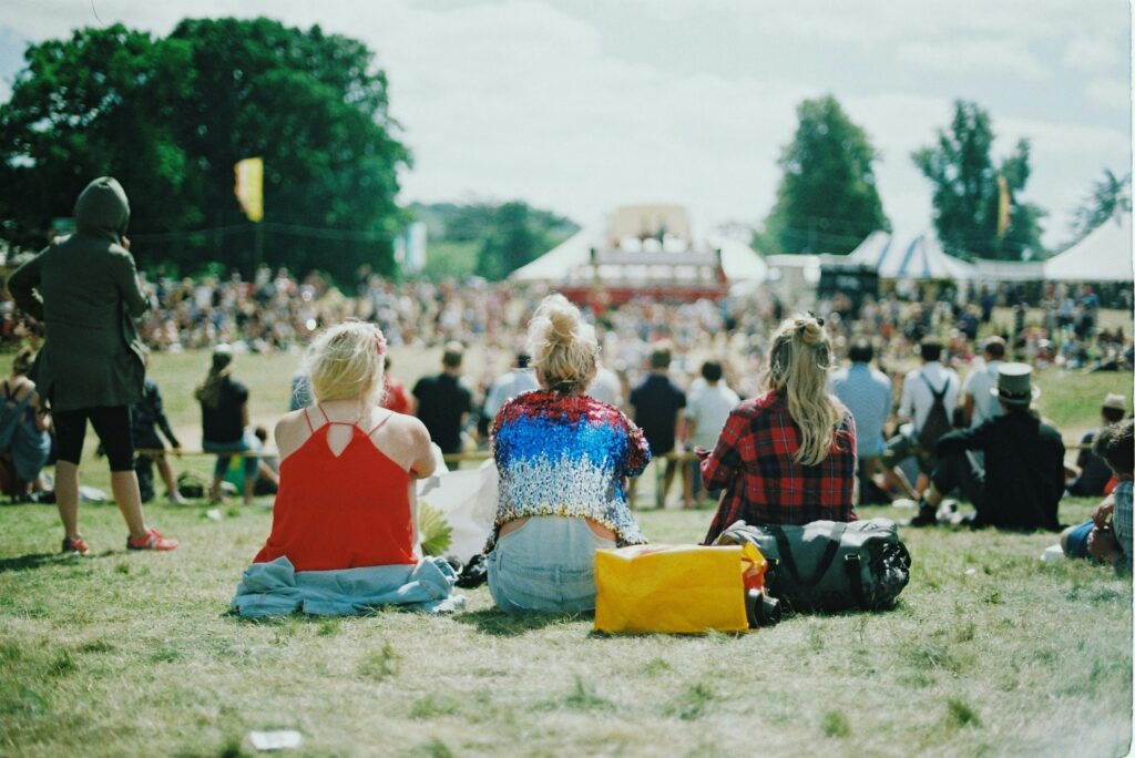 Tri osebe, ki sedijo na travi na festivalu, za njimi množica ljudi.