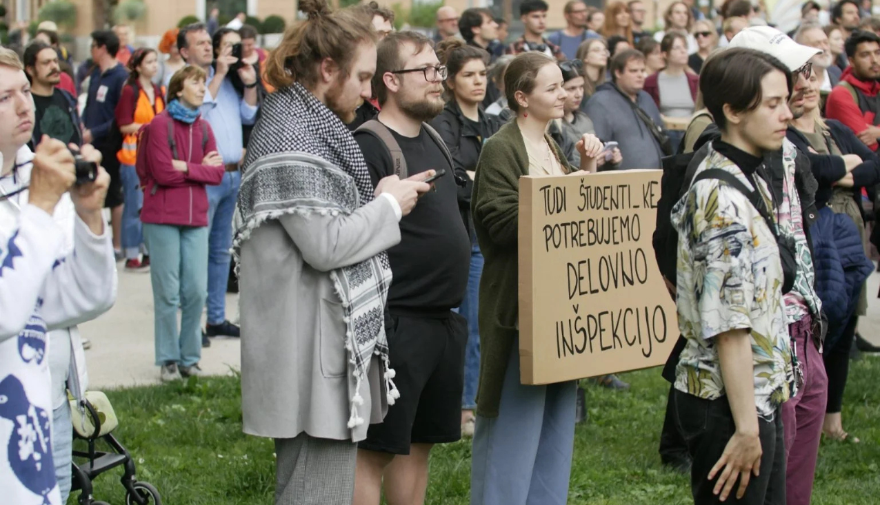 Na fotografiji stojijo mladi ljudje na protestu v parku. Mlado dekle v rokah drži transparent, na katerem piše: 'Tudi študentje potrebujemo delovno inšpekcijo'
