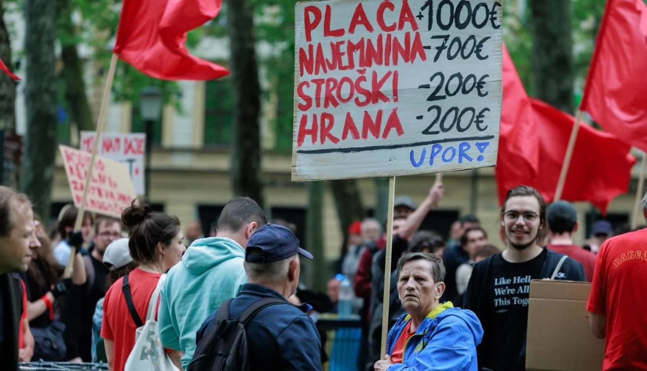 Protest ob prazniku dela. Gospa drži transparent, ki ponazarja prihodke in odhodke povprečnega državljana oziroma nekoga, ki prejema minimalno plačo.