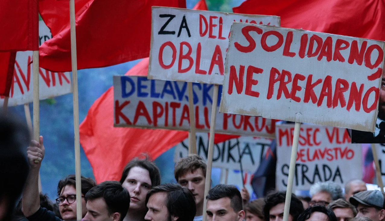 Fotografija iz protesta za 1. maj leta 2024. Prikazuje transparente, ki pozivajo k omejevanju prekarnega dela.