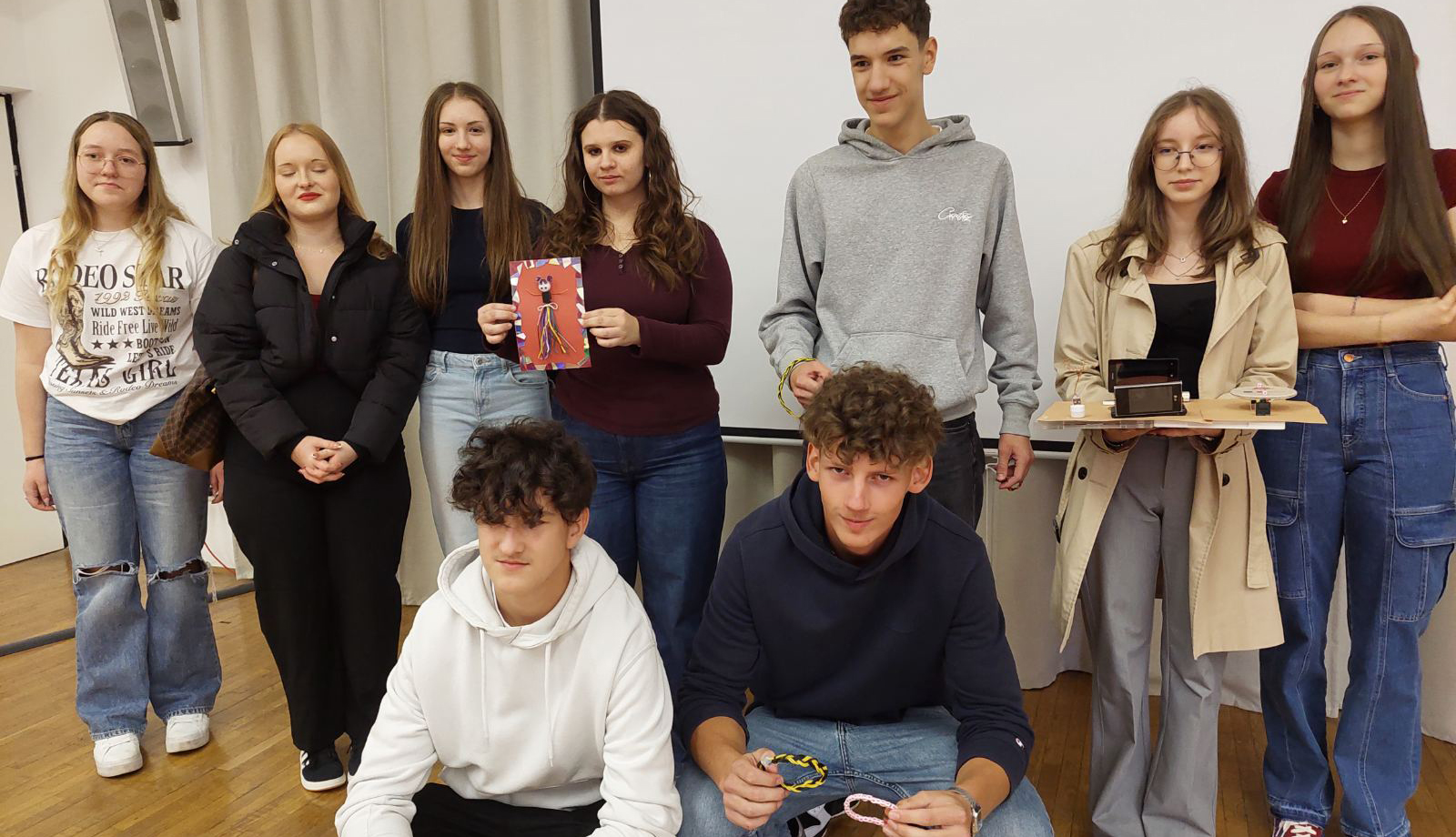 Skupinska fotografija devetih mladih v svetlem prostoru. Sedem jih stoji v zadnji vrsti, dva fanta pa klečita spredaj. Nekateri v rokah držijo izdelke, kot so barvna slika, tehnični modeli na kartonu in pletene zapestnice. Vsi so oblečeni sproščeno in gledajo v kamero.