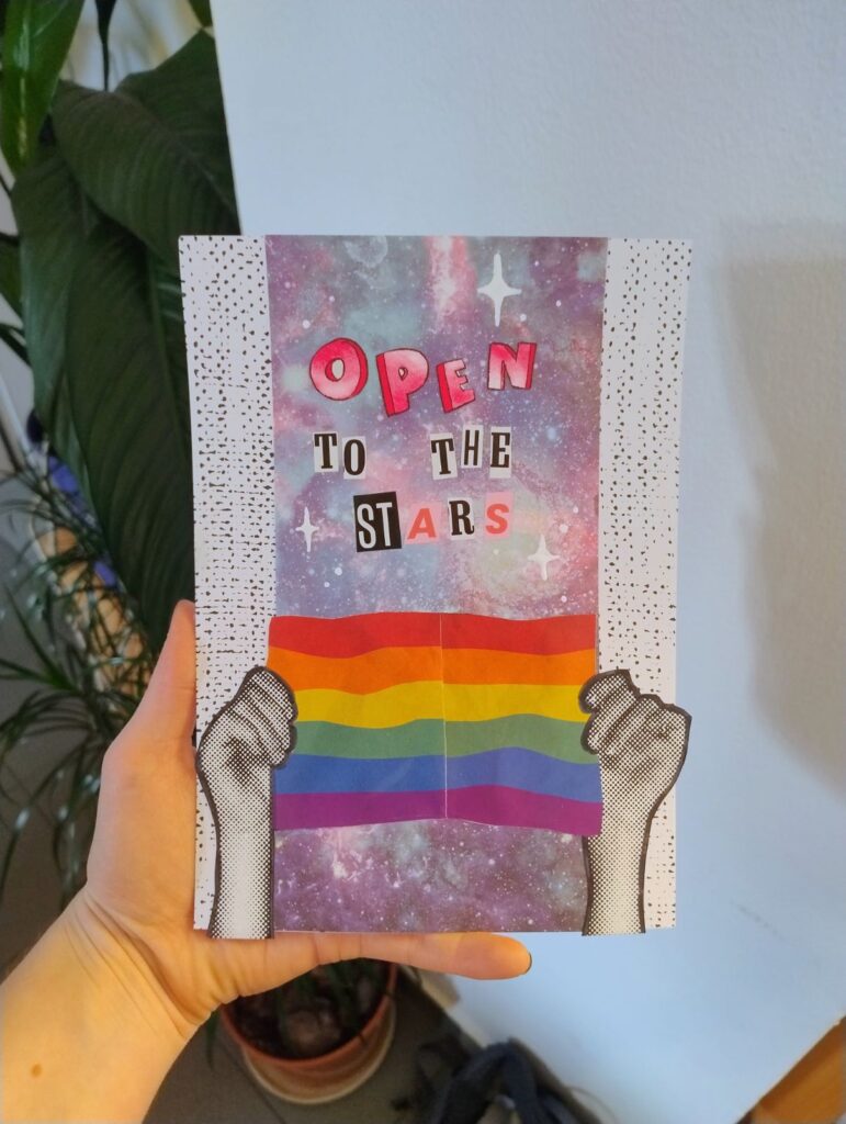 Izdelek udeleženke. Prikazuje ozadje z zvezdami in dve roki, ki držita mavrično zastavo, nad njima pa piše "Open to the stars"