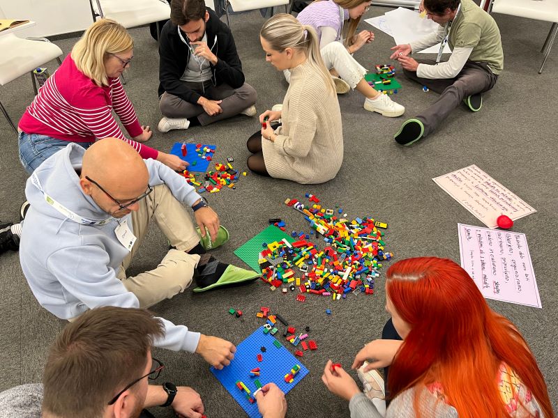 Skupina odraslih sedi na tleh v učilnici, delo v skupinah, vaja z lego kockami